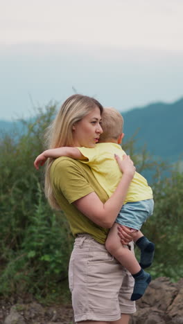 positive mother taps and strokes boy back holding son in arms on rocky hill against winding river and mountains at highland on cloudy day slow motion