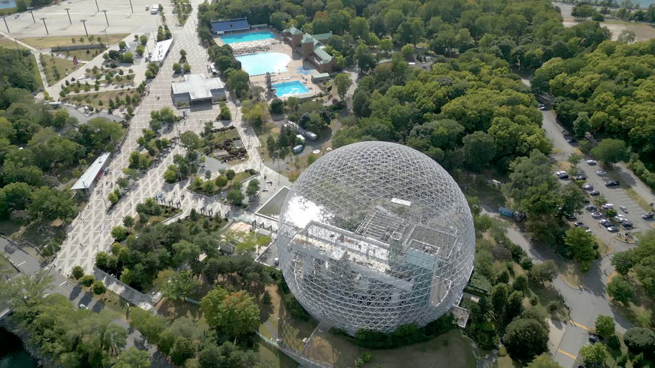 Vista aérea em ângulo alto da cúpula da Biosfera de Montreal, emoldurada por árvores e passarelas, estabelecendo a órbita