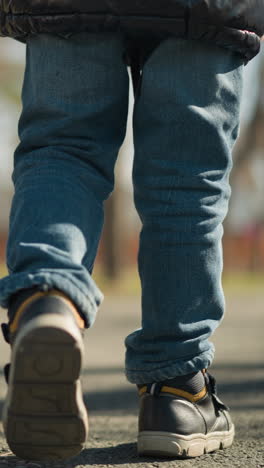 una vista de la pierna de un adulto y un niño caminando por un pavimento, el adulto lleva pantalones vaqueros y botas, mientras que el niño está vestido con una chaqueta de cuero negra, pantalones viejos y botes, con un fondo borroso