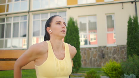 una mujer joven con auriculares realiza flexiones en un banco en un parque de la ciudad en cámara lenta. entrenando a una mujer joven en un banco con el telón de fondo de la ciudad y las casas