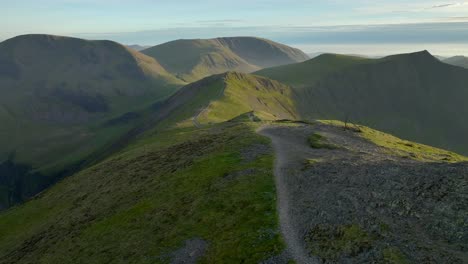 Mountain-summit-path-with-camera-rise-revealing-path-winding-down-across-ridge-lit-by-golden-hour-sun