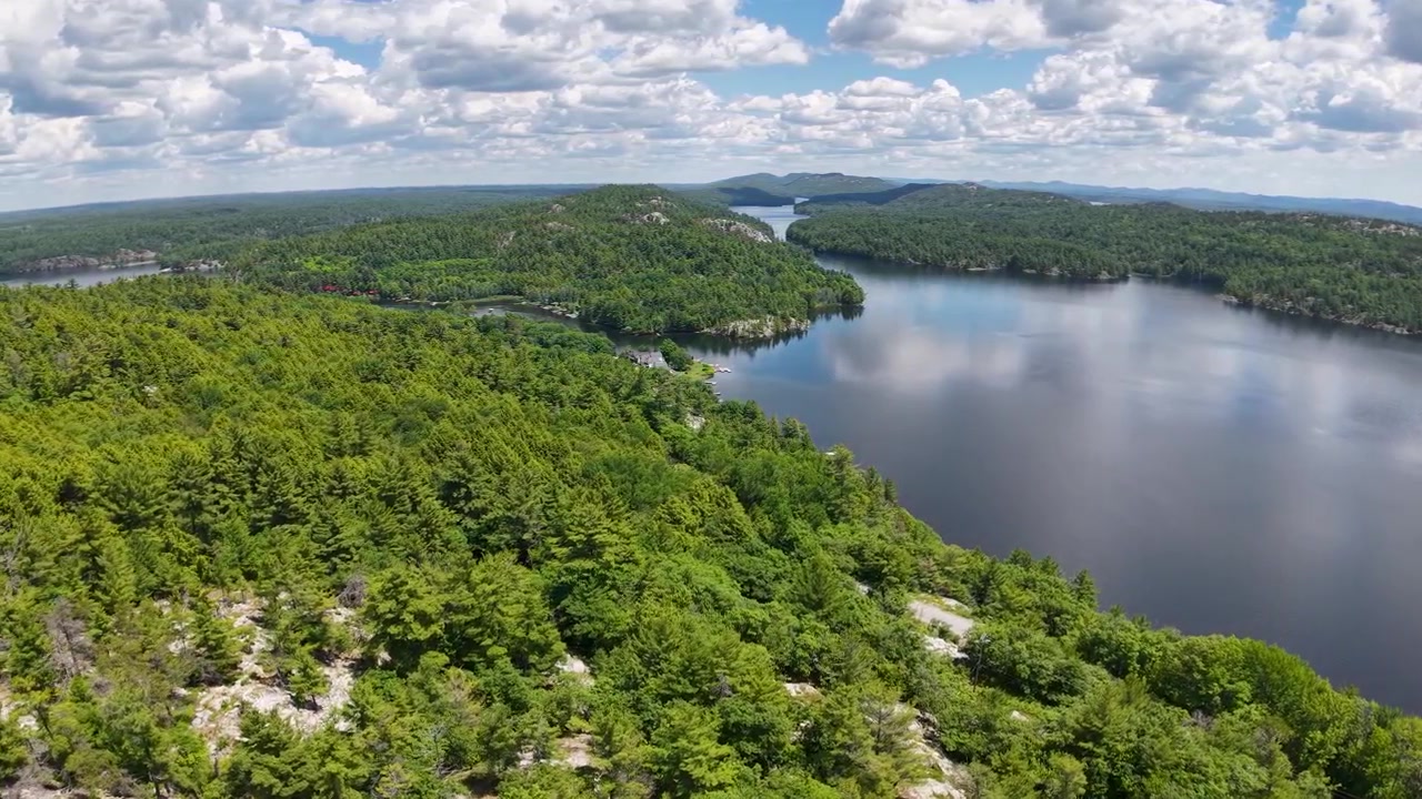 Drone rises from a rocky pine ridge to reveal a broad calm lake surrounded by continuous northern woodland near Whitefish Falls