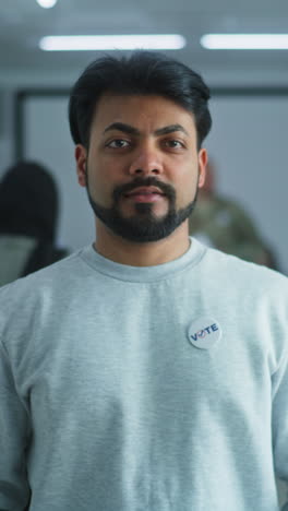 portrait of indian man, united states of america elections voter. man stands in a modern polling station, poses and looks at camera. background with voting booths. civic duty and patriotism concept.