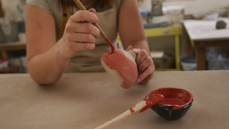 mid section of female caucasian potter wearing apron using brush to paint pot at pottery studio