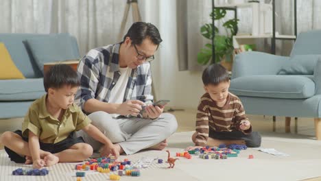 full body of asian father and sons playing the construction set colorful plastic toy brick at home. the father using smartphone, telling the kids to assemble plastic building blocks