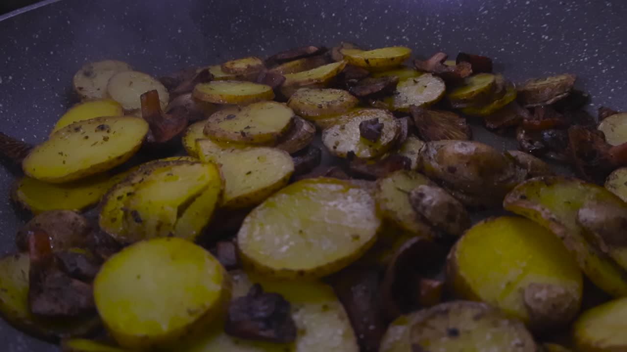 Primo piano di patate tagliate a fette e condite e funghi lattari arancioni fritti e cotti in una padella da cucina casalinga bollente al chiuso sotto la luce dei riflettori. Il cibo è condito con sale e pepe.