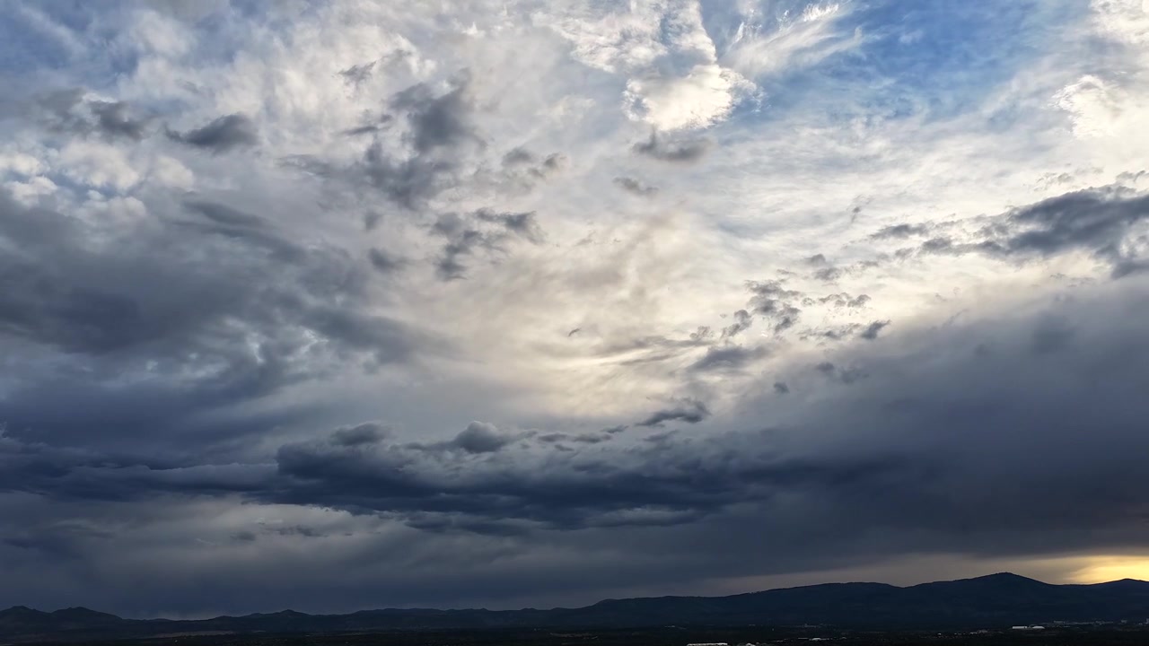Timelapse de dron 4K UHD: densas formaciones de nubes en capas dominan un cielo expansivo sobre siluetas de montañas oscuras. Un cambio atmosférico dinámico captura la llegada de fuertes sistemas de tormentas durante el anochecer