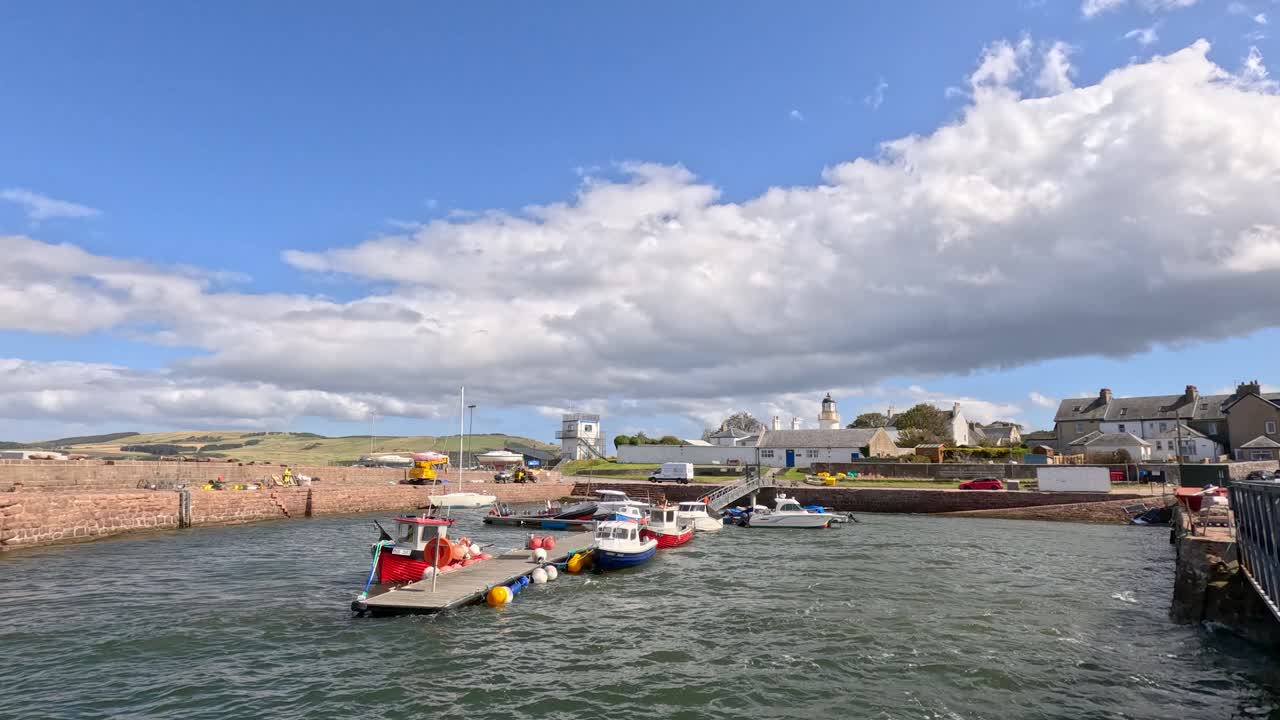 Small fishing boats maneuver into Cromarty harbor under bright daylight, with stone piers, village buildings, and dynamic clouds in the background. Wide, stable shot
