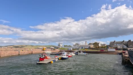 small fishing boats maneuver into cromarty harbor under bright daylight, with stone piers, village buildings, and dynamic clouds in the background. wide, stable shot
