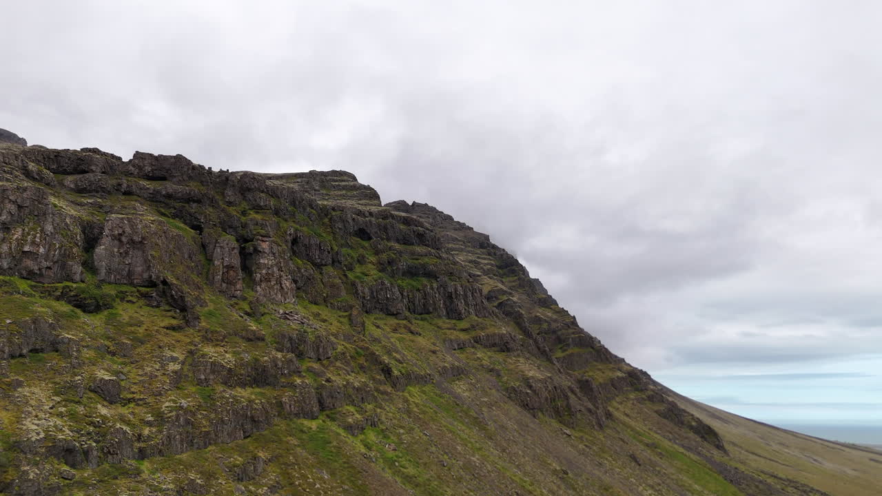 Aerial view of steep mountain cliffs in Sveitarfélagið Hornafjörður, showing layered rock formations, glacial valley textures, and sweeping rugged terrain under an overcast sky
