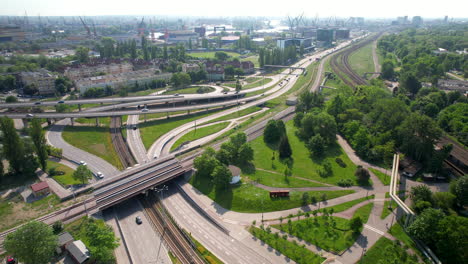 Aerial-flyover-intersection-and-road-with-bridges-and-traffic-in-Gdansk-during-sunny-day---Industrial-harbor-cranes-in-background