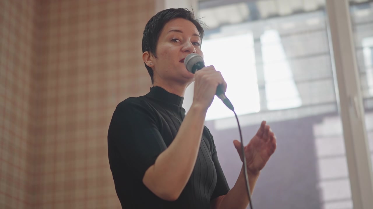 Cantante che si esercita con il microfono accanto alla finestra, prova intima in aula, sfondo con carta da parati a quadri, espressione concentrata, esercizio di respirazione, dimostrazione di coaching, silhouette in controluce