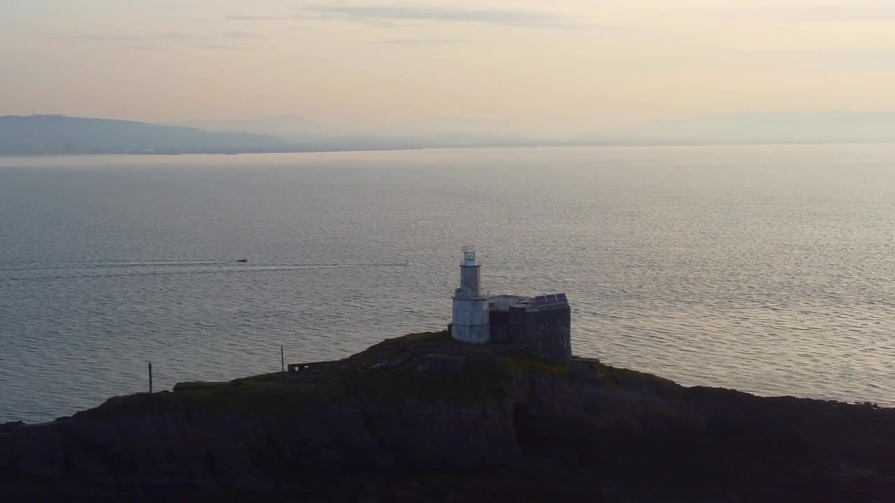 Ripresa aerea laterale di barche che si dirigono verso il mare con il faro di Mumbles all'alba. Riflesso dorato del sole sull'oceano con nuvole dinamiche. Perfetto per il turismo, documentari di viaggio. Galles, Regno Unito. 4K.