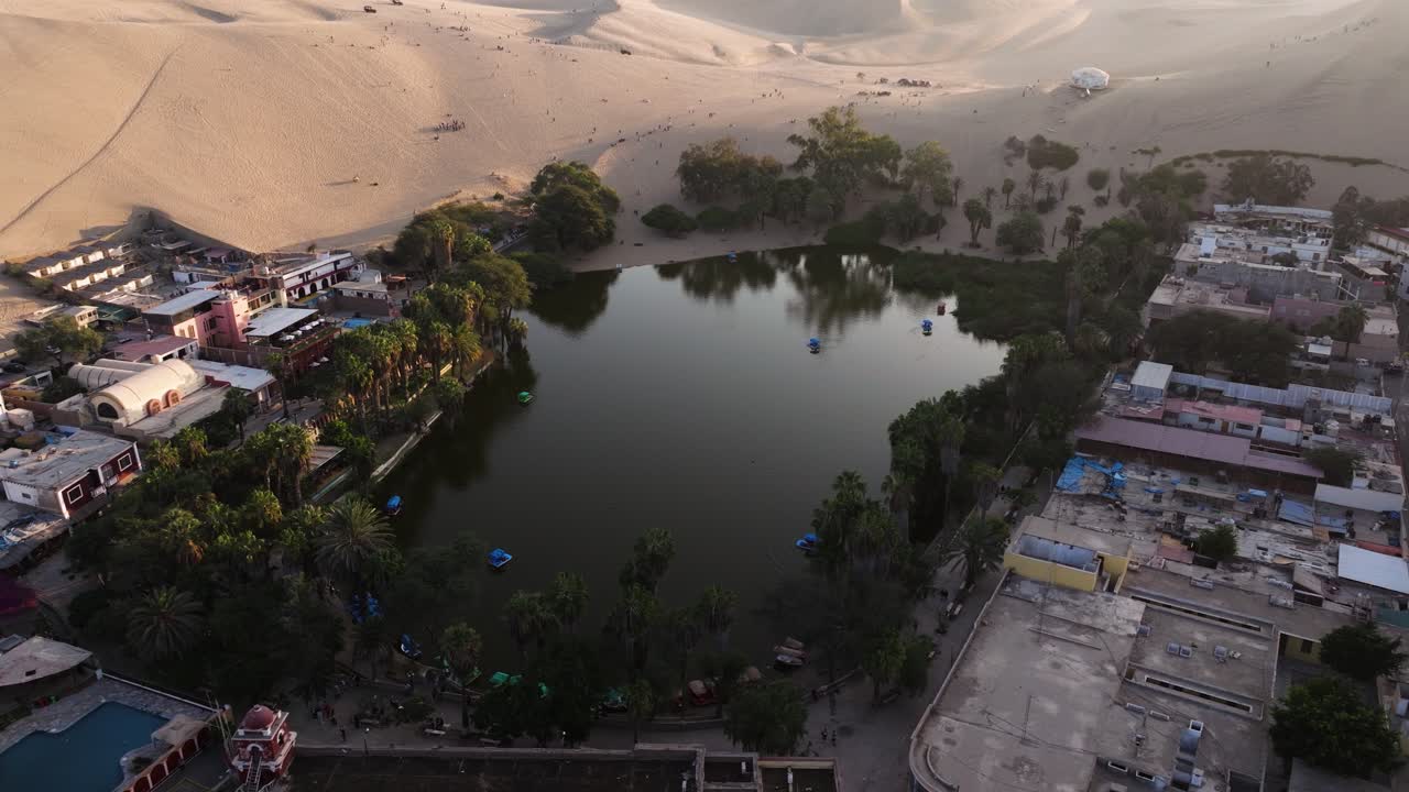 Aerial angled orbit of Huacachina oasis, layered sand textures surrounding ponds and buildings, establish