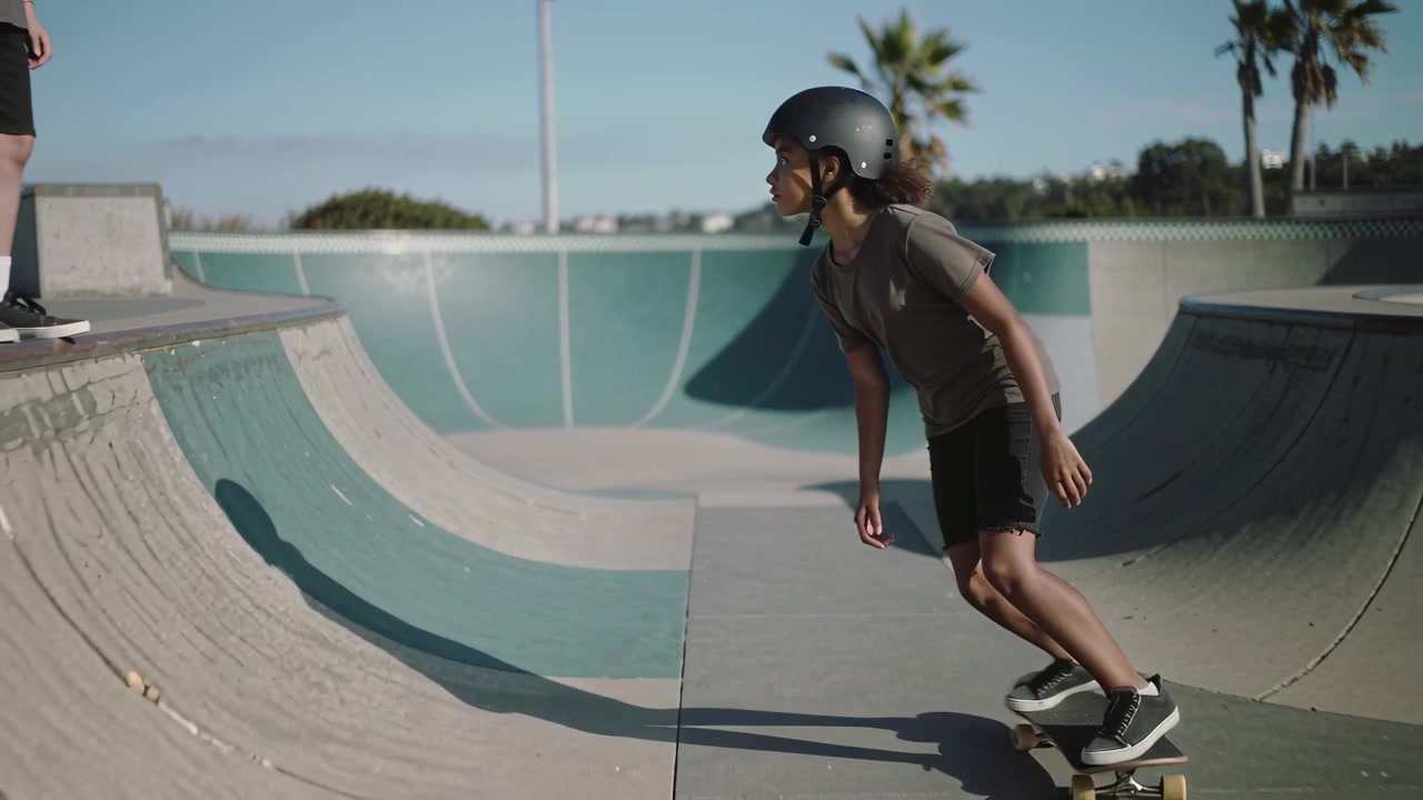 Dropping teen skateboarder wearing helmet pushing into skate bowl, carving and looking toward peers