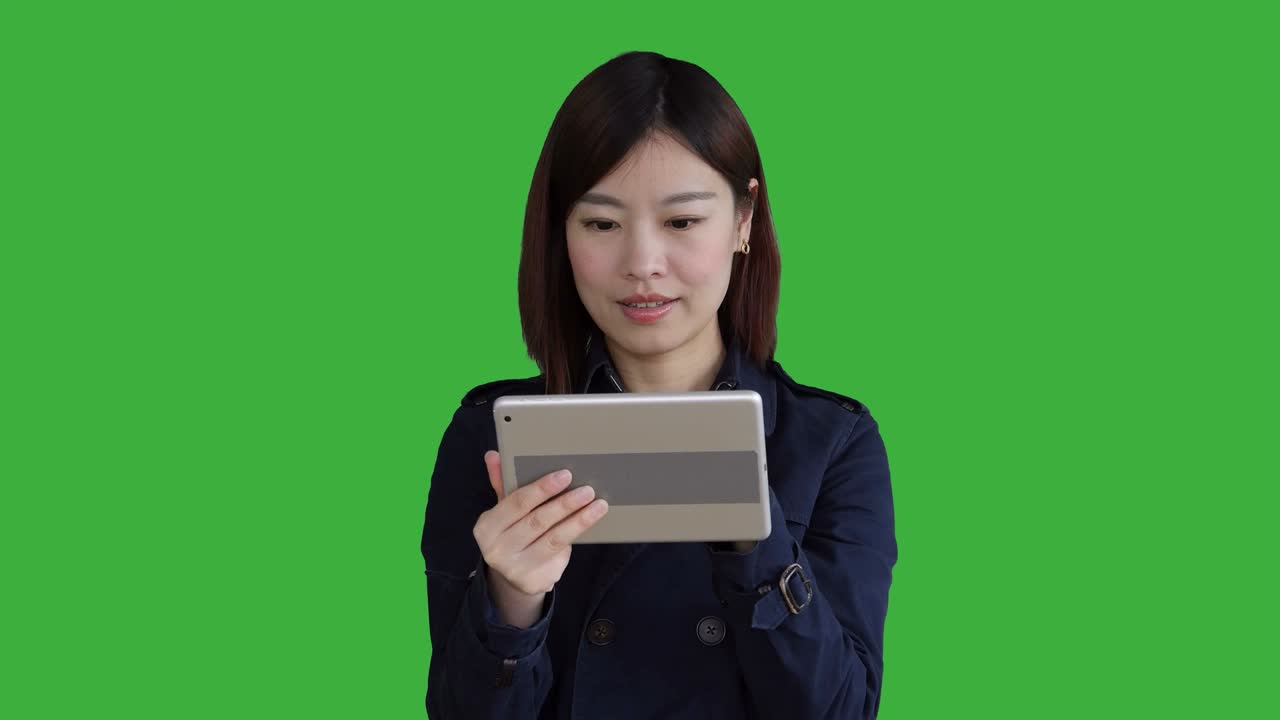 푸른 코트를 입은 젊은 아시아 여성, 스마트폰, 디지털 태블릿을 사용하고 카메라를 보고, 크로마 키를 위해 녹색 화면 앞에 서있는 여성.