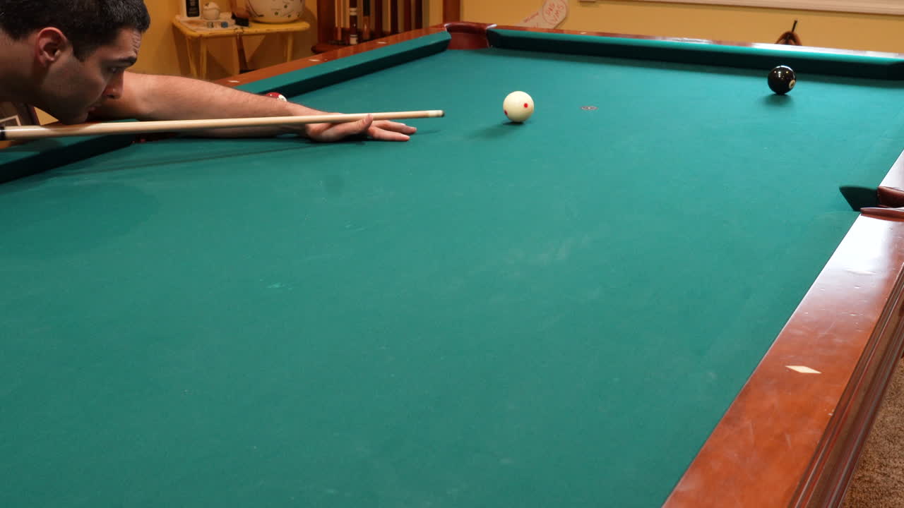 Man Playing 8 Ball Pool Shoots Solid Black Eight Ball into Corner Pocket to Win the Game on a Brunswick Table with Green Felt in Basement of Home, Open Bridge Hand and Wooden Cue Stick, Wide Angle