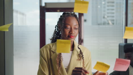 african american business woman write tasks creative ideas on sticky notes on glass board female corporate leader planning project on post it sticky notes organize work on stickers