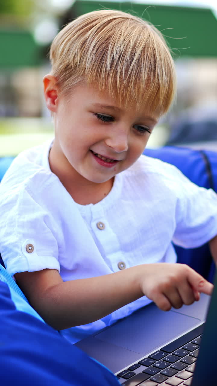 Caucasian six-year old with laptop outdoors. Peaceful boy in bean bag chair outdoors playing computer. Blurred backdrop. Vertical video