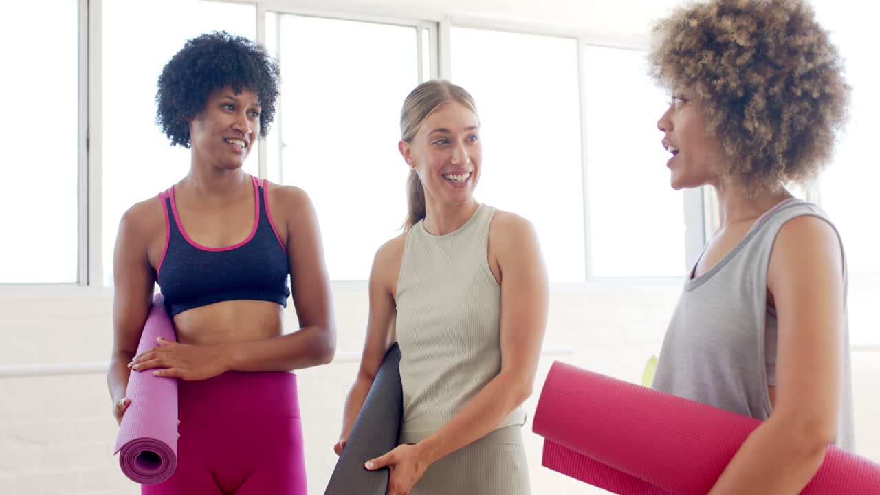 tres mujeres están charlando en un estudio de yoga brillante, sosteniendo esteras