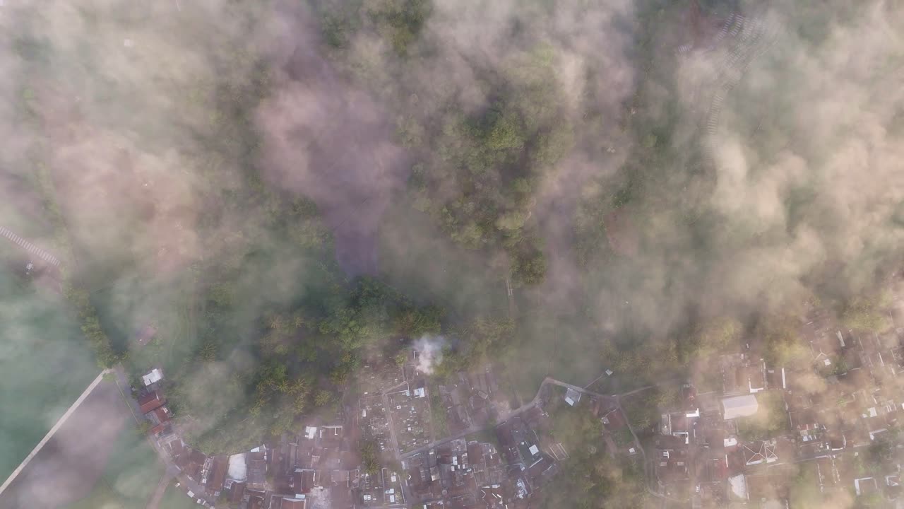 Overhead drone view showing foggy morning over a countryside village with rooftops and forest canopy under the mist. Indonesia tropical rural landscape