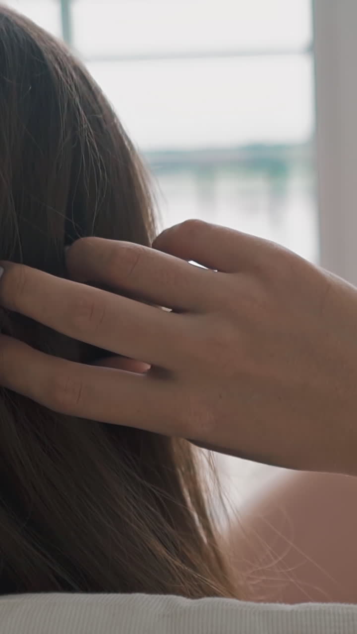 Young woman touches long brunette hair by hand resting in armchair with pillows against large blurry window in light room extreme close backside view