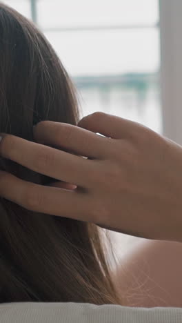 young woman touches long brunette hair by hand resting in armchair with pillows against large blurry window in light room extreme close backside view
