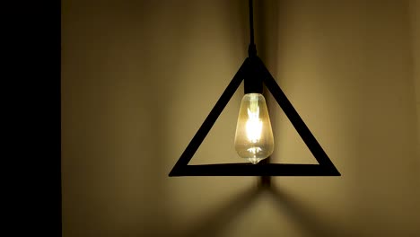 Closeup-of-a-triangle-lamp-hanging-on-a-wall-and-shining-with-golden-light-on-the-wall-at-darkness,-static-and-copy-space