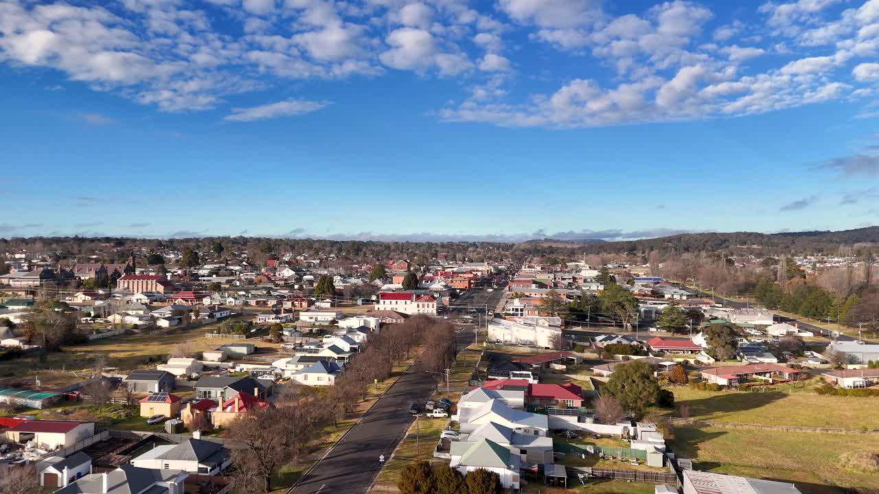Drone camera glides forward above a quiet residential street in a small Australian town, revealing houses, buildings, and clear blue sky in bright daylight