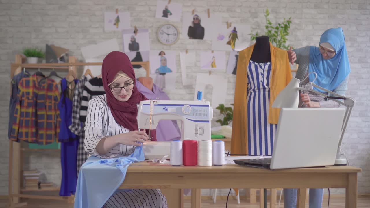 Perancang busana muslim muda mengenakan jilbab nasional sedang bekerja dengan mesin jahit di studio.