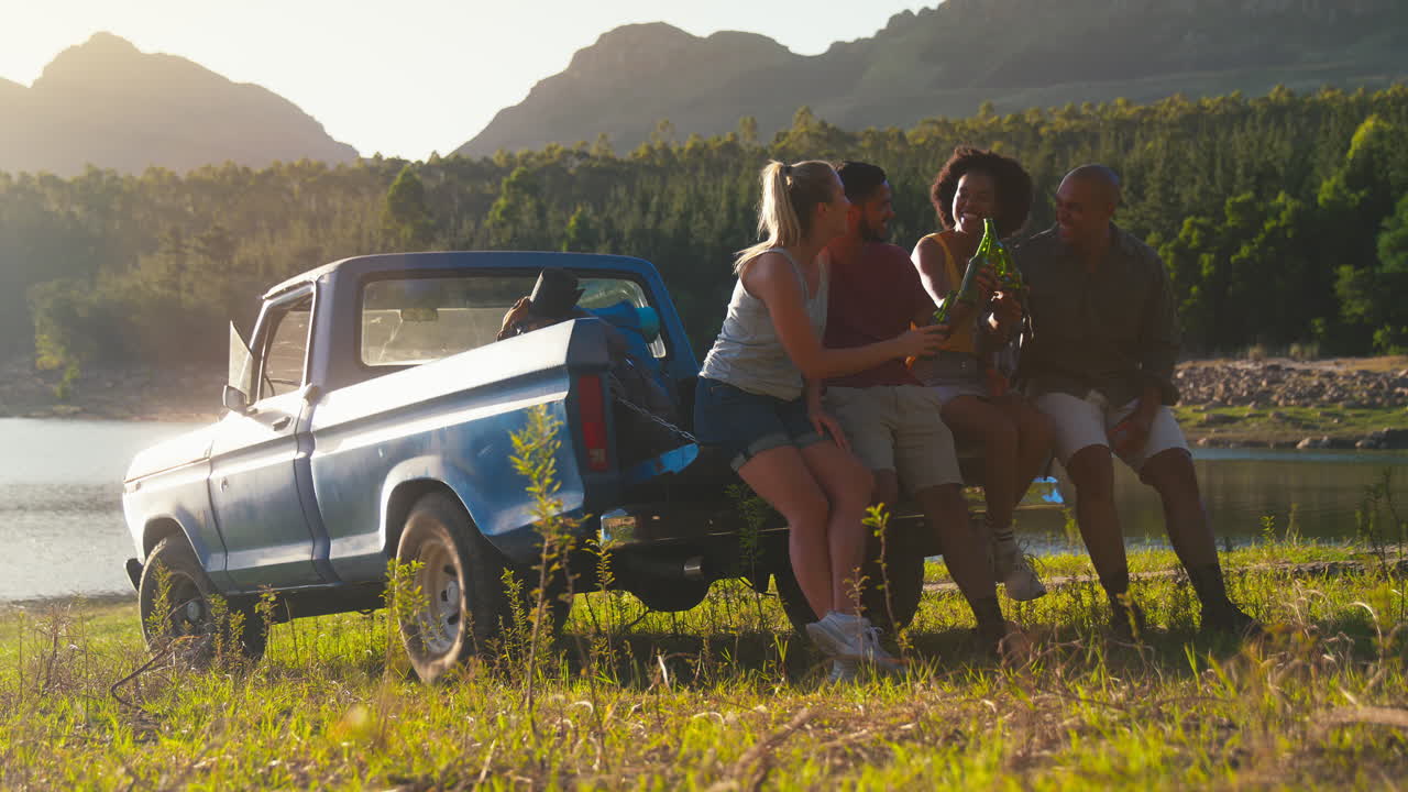 배 을 들고 있는 친구 들 이 호수 를 따라 도로 여행 을 하는 트럭 의 뒷문 에 앉아 맥주 를 마시고 있다