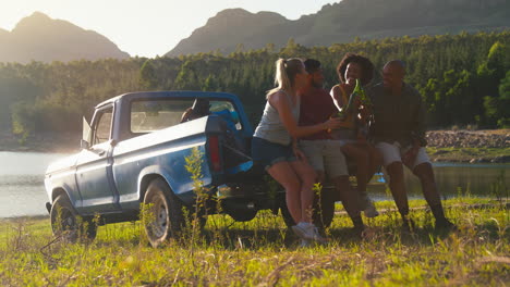 Friends-With-Backpacks-Sitting-On-Tailgate-Of-Pick-Up-Truck-On-Road-Trip-By-Lake-Drinking-Beer