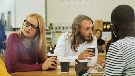 Camera-focuses-on-a-multiethnic-group-of-friends-through-the-window-talking-while-drinking-coffe-sitting-at-a-table-in-a-cafe