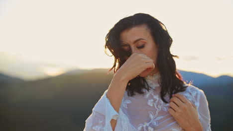 sad thoughtful lady in white dress touches her dark hair gently. woman posing in the mountains against bright setting sun. close up.