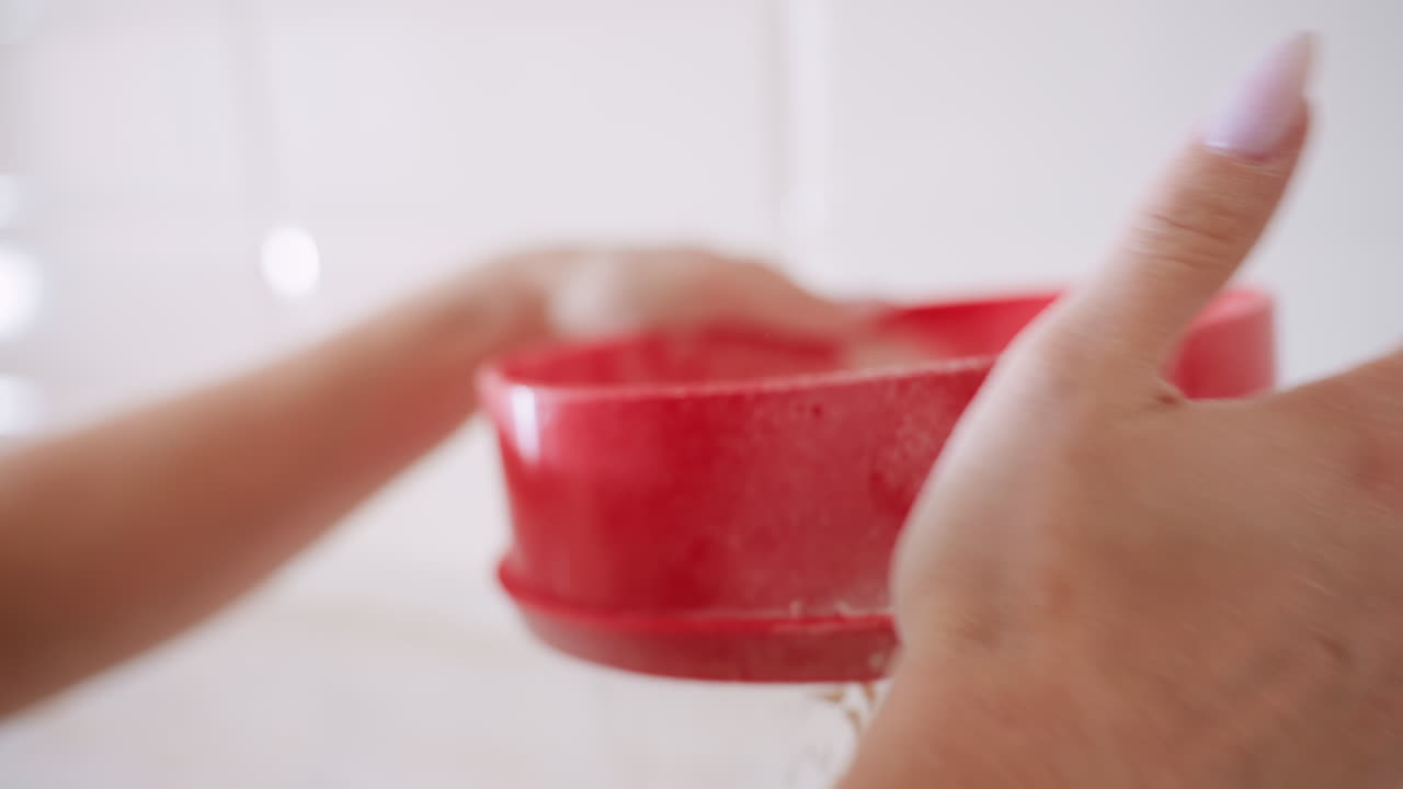 Close up of baker hand filtering flour with red sieve into bowl during baking preparation, hands in focus as fine powder falls smoothly creating texture and detail for homemade recipe
