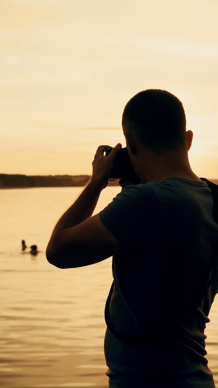man with a camera in his hands makes a photo of the river on the background of sunset. Vertical video