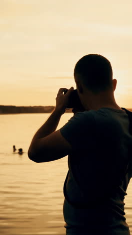 man with a camera in his hands makes a photo of the river on the background of sunset. vertical video