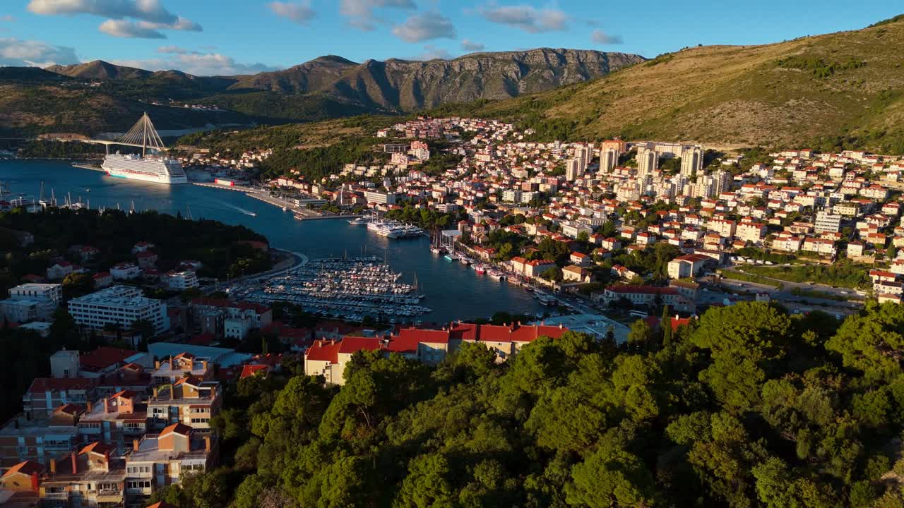 Aerial drone captures a sweeping landscape of Dubrovnik’s coastal town and cruise ship port, with majestic mountains rising in the background, as the warm golden sunset bathes the city and sea