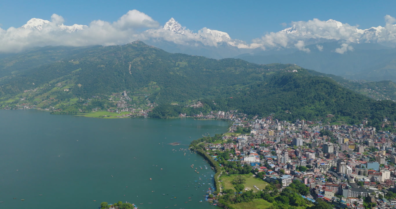 Aerial shot of Phewa Lake Nepal showing calm blue water surrounded by green hills and a lakeside city with Annapurna and Machhapuchhre peaks rising in Pokhara.