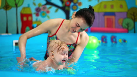 funny baby boy is crying when coach splashes water on him. infant kid is taught to swim.