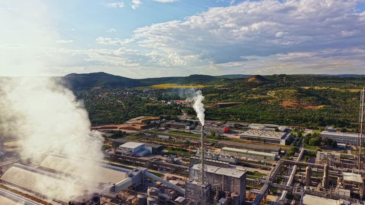 Fabbrica con fumo che sale nel cielo di giorno, vicino a colline verdi e una piccola città sullo sfondo