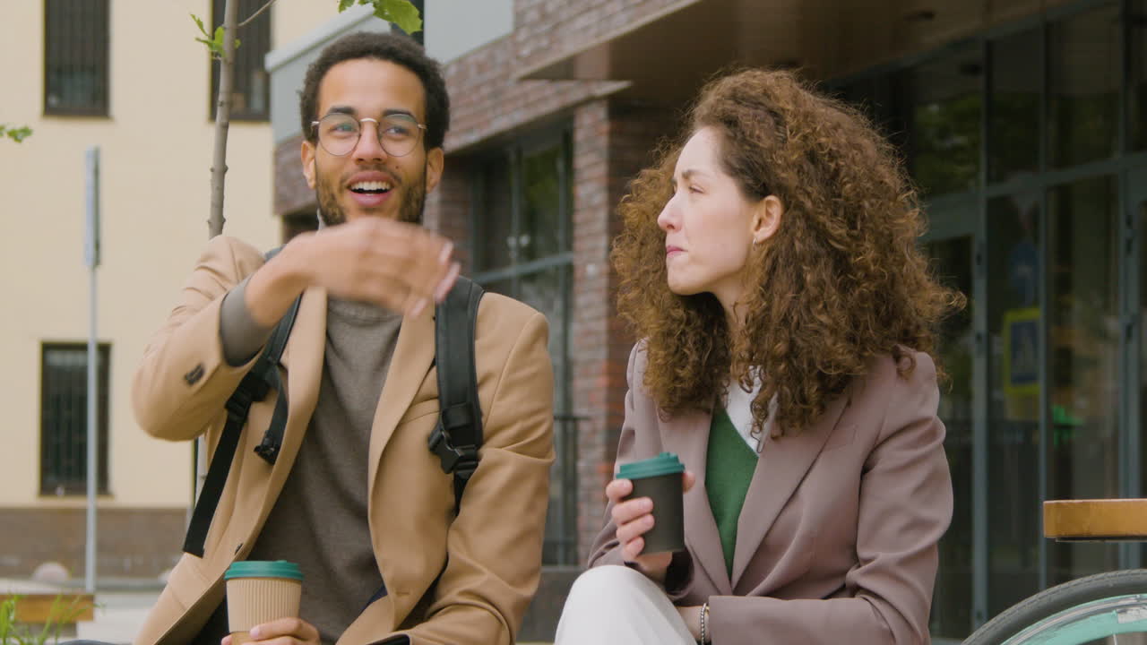 Young American Man And Woman In Formal Clothes Holding Takeaway Coffee And Talking While Sitting On A Bench In The City