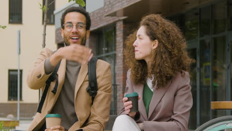 Young-American-Man-And-Woman-In-Formal-Clothes-Holding-Takeaway-Coffee-And-Talking-While-Sitting-On-A-Bench-In-The-City