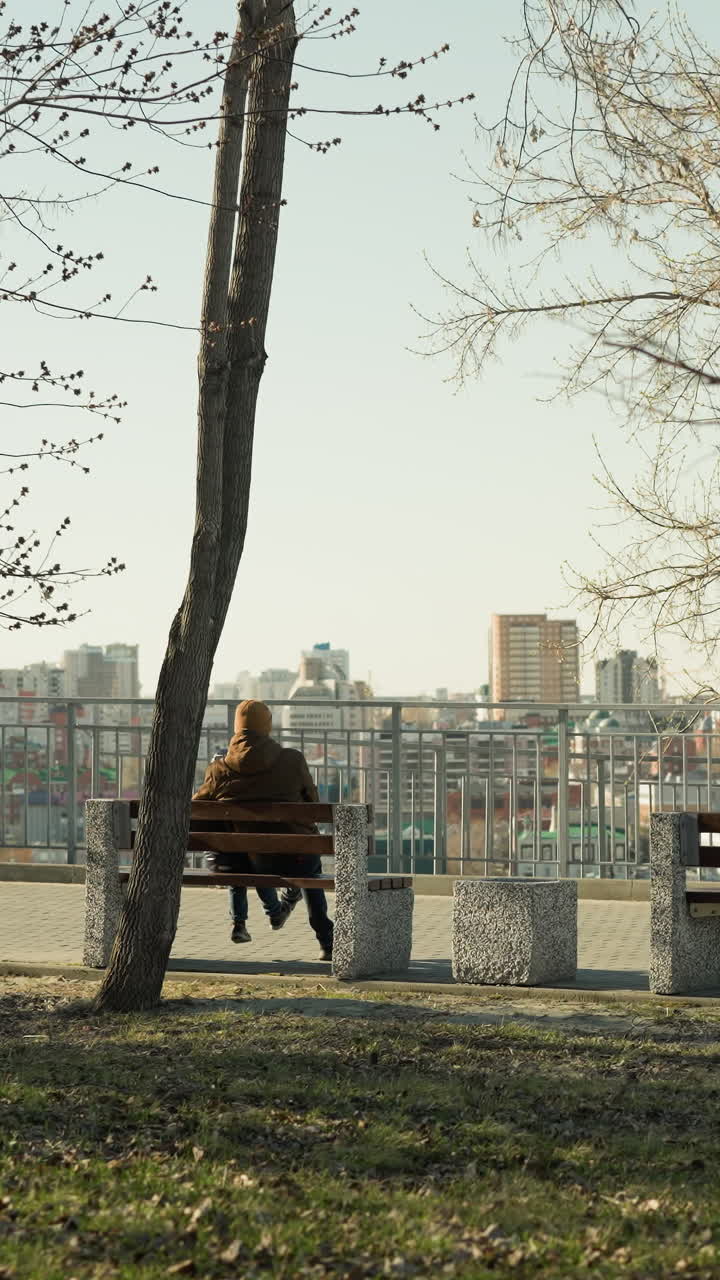 padre e hijo sentados juntos en un banco, mirando el paisaje de la ciudad desde un parque al aire libre, el padre tiene las piernas cruzadas mientras el niño golpea suavemente su pierna