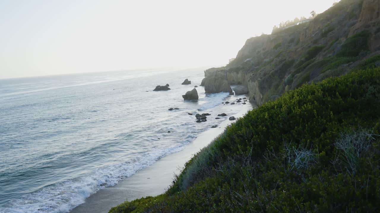A coastal cliff view overlooking the ocean, dotted with rocks and gentle waves lapping against the beach during early morning light