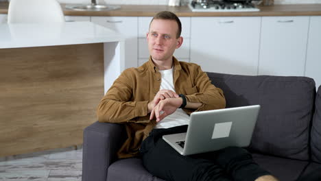 Male freelancer using laptop while laying on couch. Young man looking at his watch and talking to someone while working on a laptop at home.