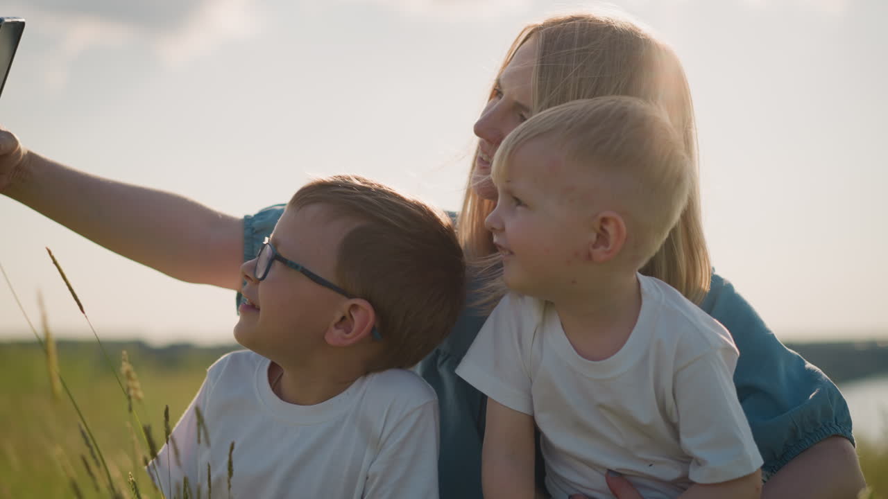 A woman in a blue dress sits on the grass, smiling as she takes a picture with a smartphone. Her two sons, both wearing white shirts, joyfully join her in capturing this sunny outdoor memory together