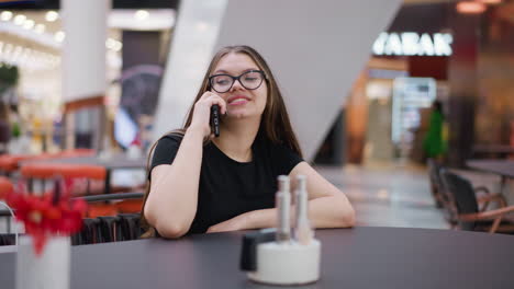 young girl with glasses on phone call, glancing to right, seated in mall food court, with blurred background featuring modern architecture and passersby