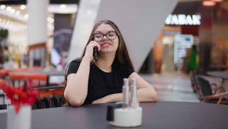 joven con gafas en una llamada telefónica, mirando a la derecha, sentada en el patio de comidas del centro comercial, con un fondo borroso con arquitectura moderna y transeúntes