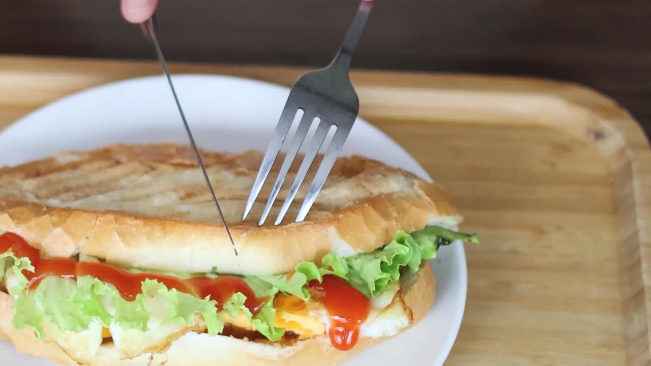 A sandwich on a plate being sliced with a fork and knife on a wooden board.
